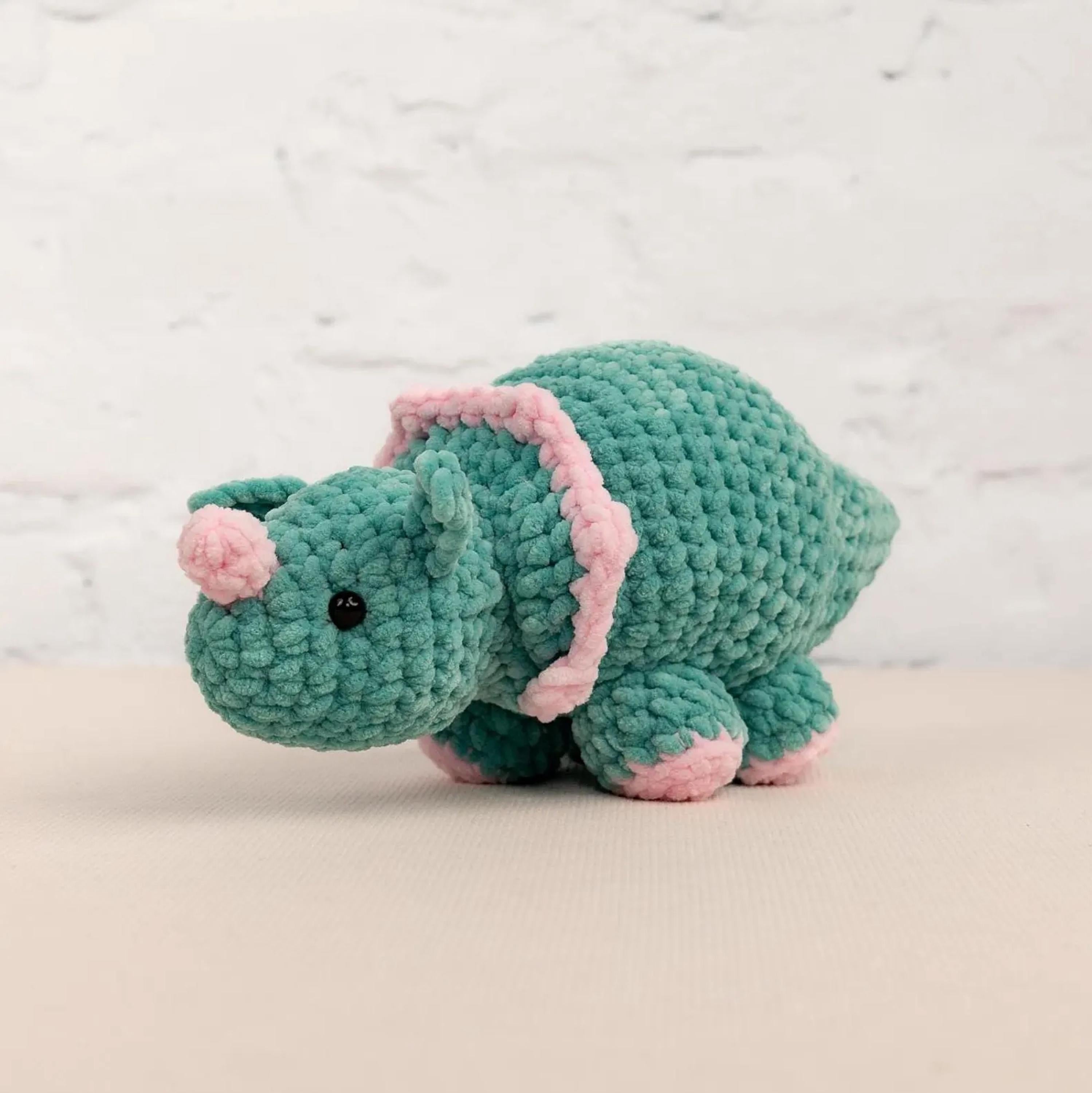 A crocheted green and pink stuffed animal toy is sitting on a white surface against a white brick wall background.